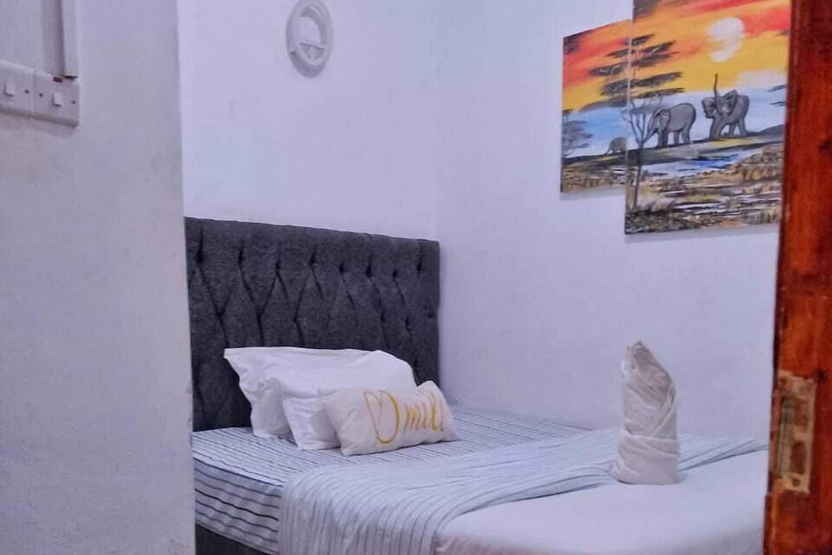 Un dormitorio con cabecera gris tapizada, cama con ropa de cama blanca y manta a rayas, alfombra roja y un cuadro de elefantes en la pared.