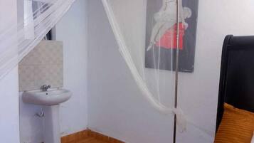 Un dormitorio con cama, mesita de noche, ventana con cortina y baño con lavamanos y espejo.
