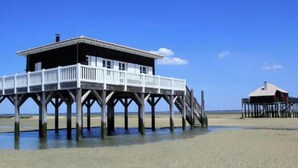 Un muelle de madera con una casa sobre pilotes, una edificación separada a la derecha y un cielo despejado.