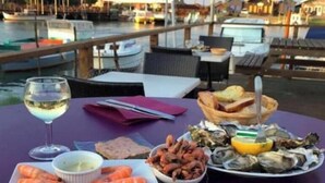 Una mesa de comedor con platos de mariscos, una copa de vino blanco y vistas a un puerto deportivo con barcos y edificios.
