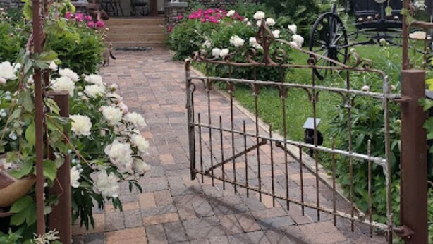 A brick pathway leading to a house with a porch, surrounded by lush greenery and a decorative metal archway. - Relax in our Fish Haven Room in the Lower Lodge at The Cobblestone Bed & Breakfast (Birchwood)