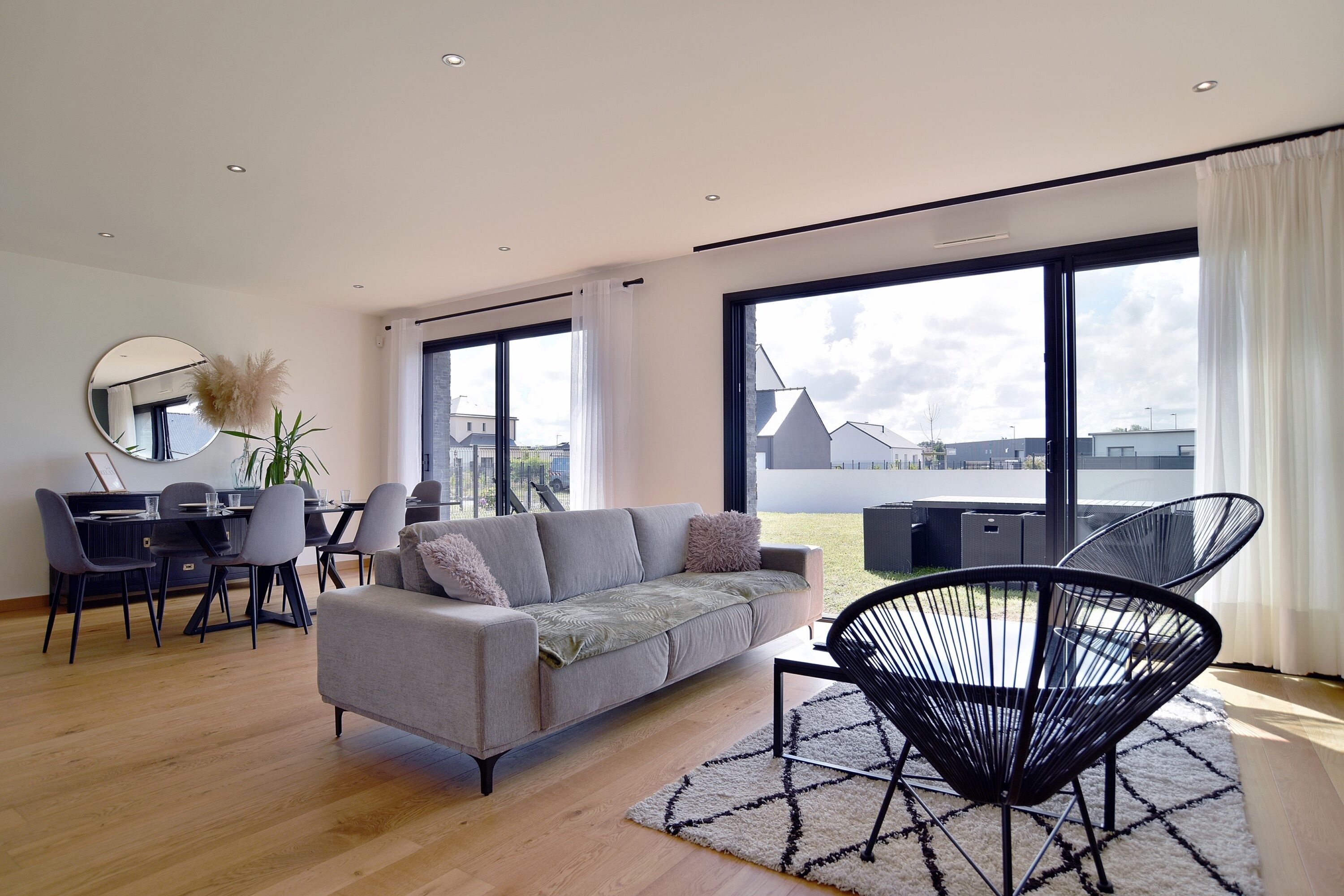 A modern living room with a grey sofa, black chair, dining table, and large windows offering a view of buildings.
