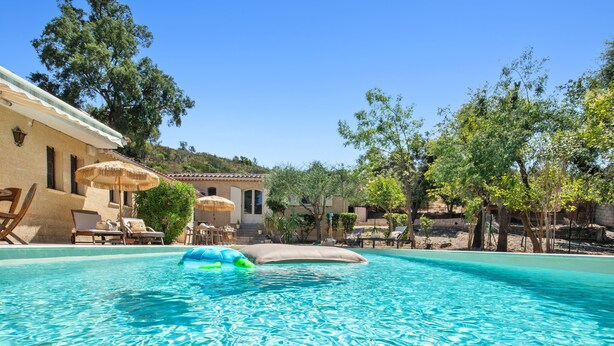 A clear blue pool with lounge chairs and umbrellas, surrounded by trees and a building with a tiled roof. - Chez Nous Baraille - Appt climatisé avec piscine (Puget-sur-Argens)