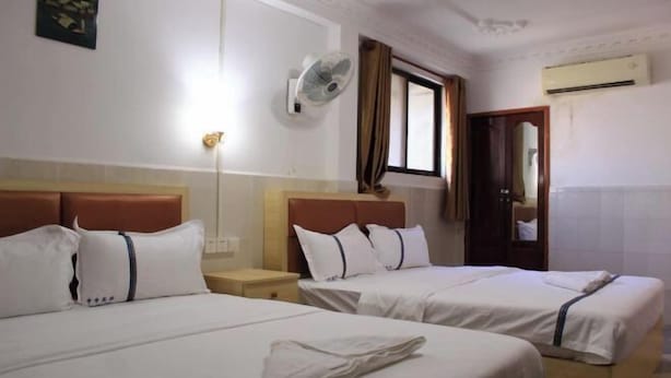 A hotel room with two beds, a wall-mounted fan, an air conditioning unit, and a window with curtains.