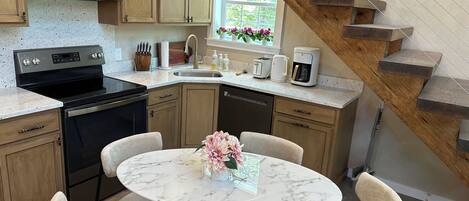 Una cocina moderna con microondas integrado, estufa y lavavajillas. Hay una zona de comedor con una mesa de mármol y sillas, y una escalera de madera que conduce a otro nivel.