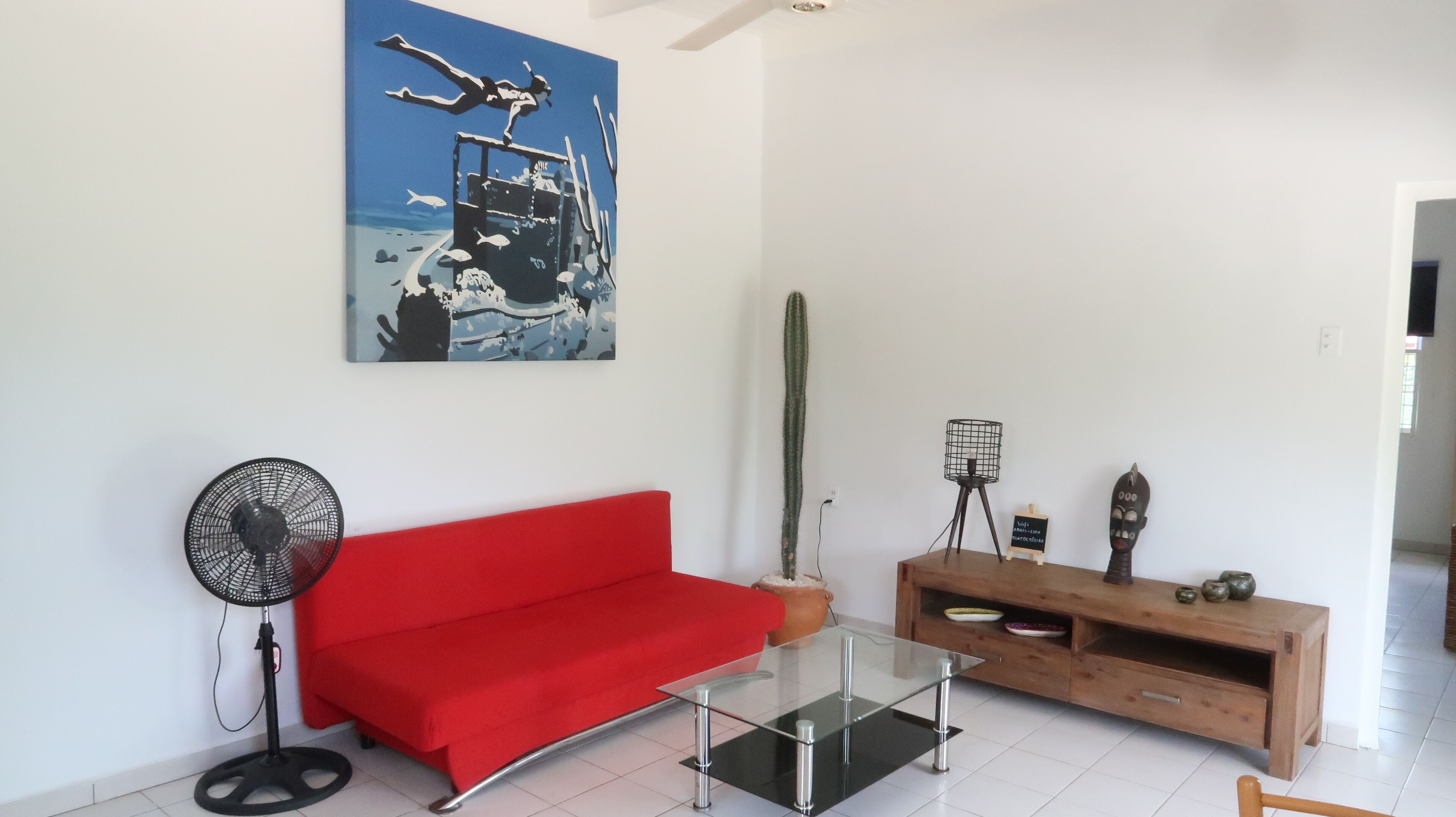Ein Wohnzimmer mit einem roten Sofa, einem Glastisch, einem Kaktus, einem Wandventilator und einem Bild von einem Taucher.