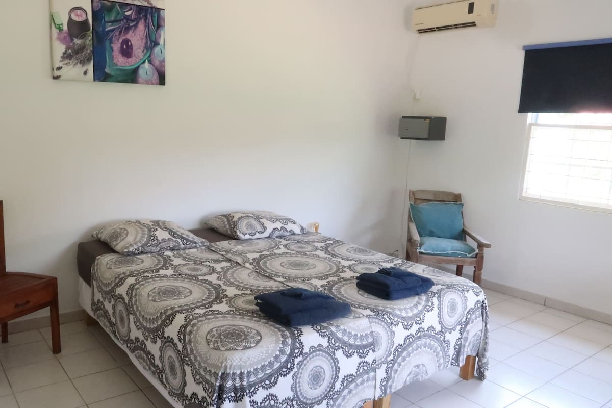 A bedroom with a bed, a chair, a small table, a wall-mounted air conditioner, and a window with blinds.