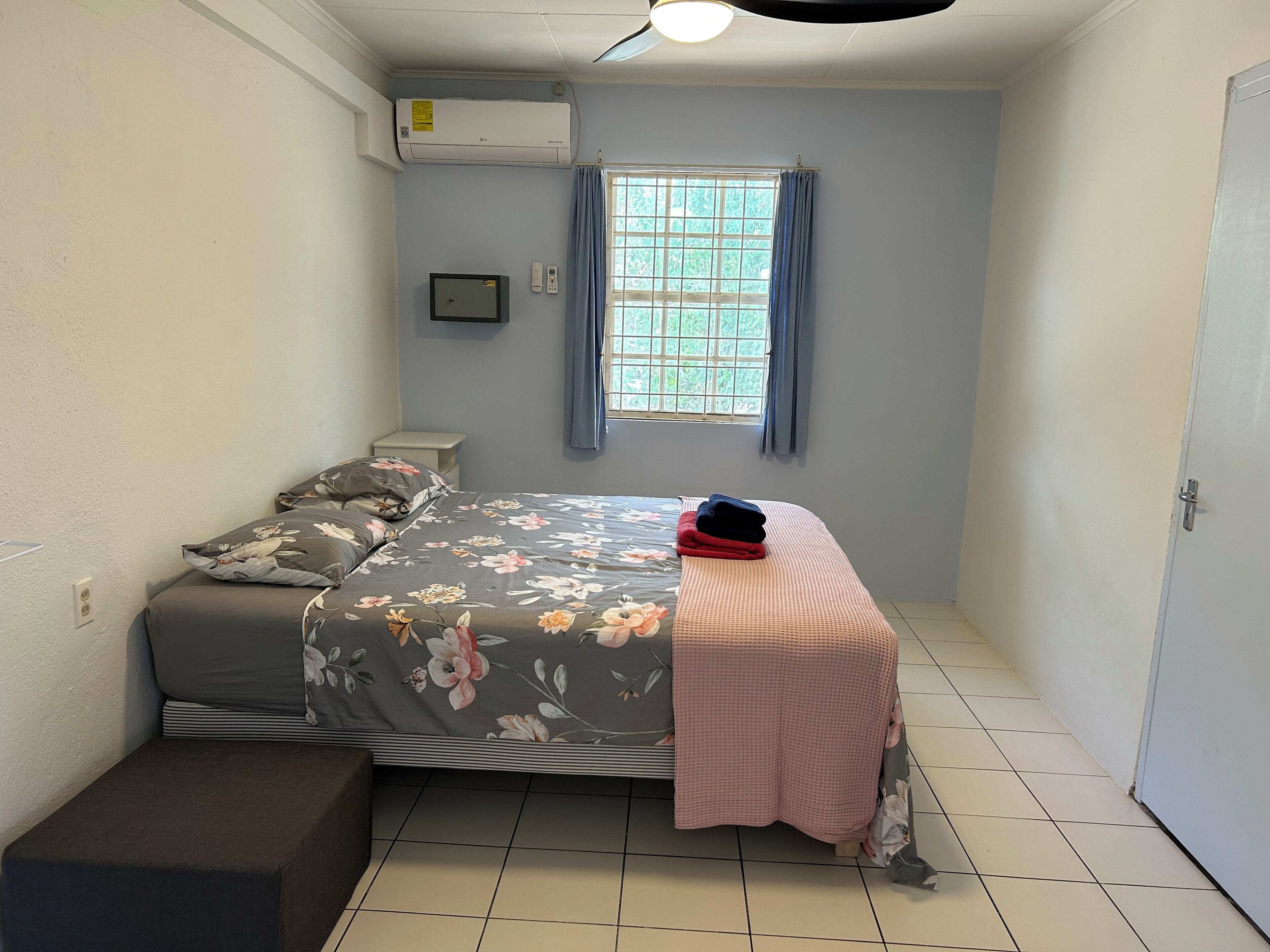 Een kleine eenpersoonskamer met raam, nachtkastje en een wandairconditioner.
