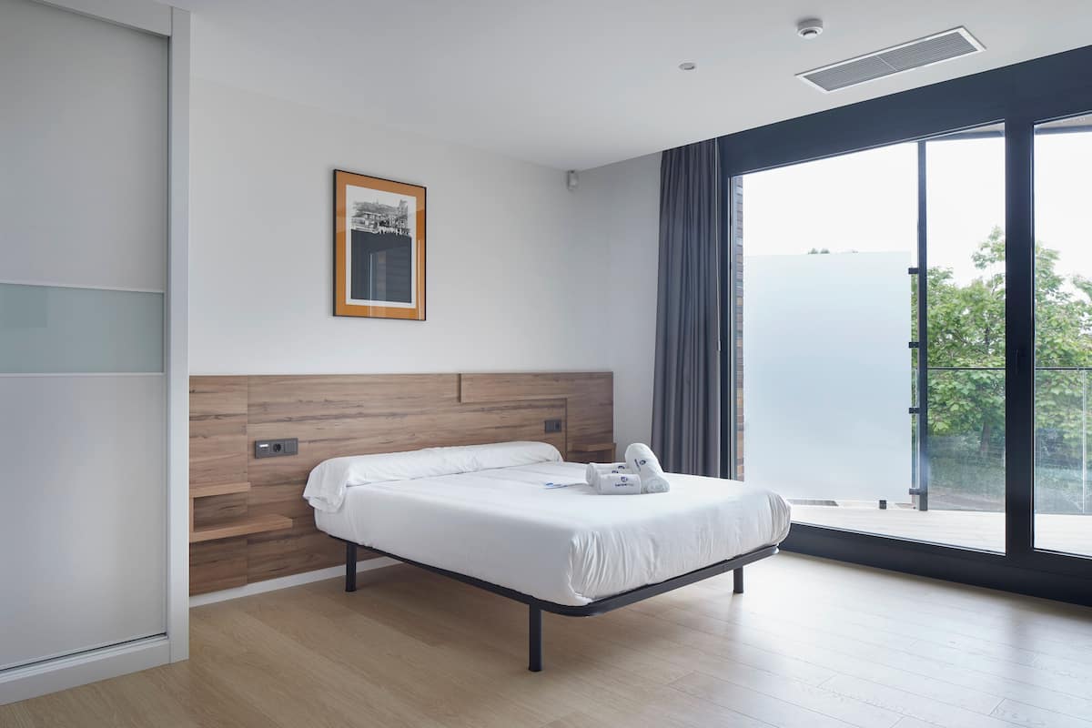 A modern hotel room with a wooden headboard, a large window with a view, and a neatly made bed with towels.