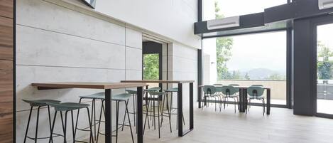 A modern interior with a long table and high stools, large windows offering a view of greenery and hills.