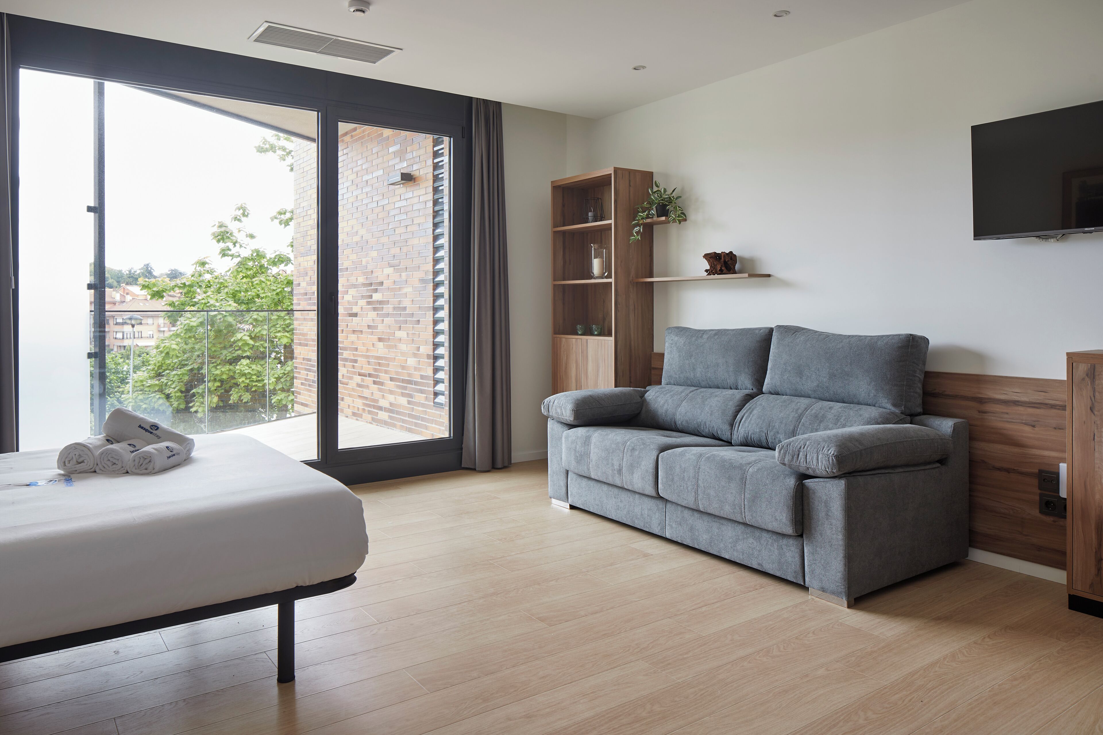 A modern bedroom with a bed, grey sofa, wooden shelves, and a TV.