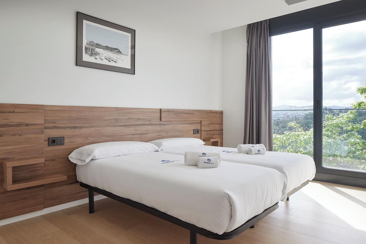 A modern hotel room with a large bed, a wooden headboard, a framed picture on the wall, and a view of greenery through the window.