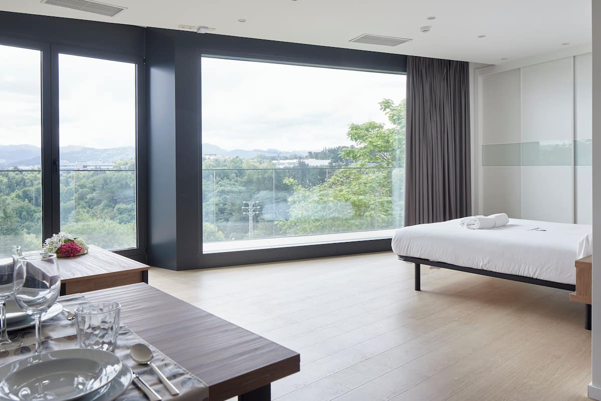 A modern hotel room with a large window, a bed, a dining area with a table set for two, and a view of the outdoors.