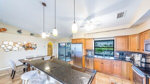 A modern kitchen with a central island, wooden cabinets, stainless steel appliances, pendant lighting, and a view through the window.
