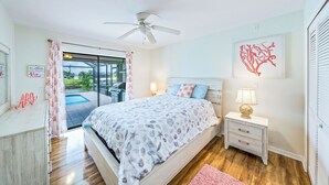 A bedroom with a bed, a nightstand, a ceiling fan, a window with curtains, and a view of the outdoors.