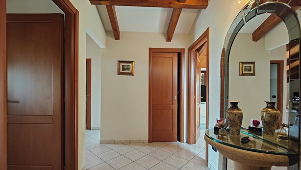 Um corredor com portas de madeira, piso de azulejos e uma mesa com tampo de vidro adornada com objetos decorativos.