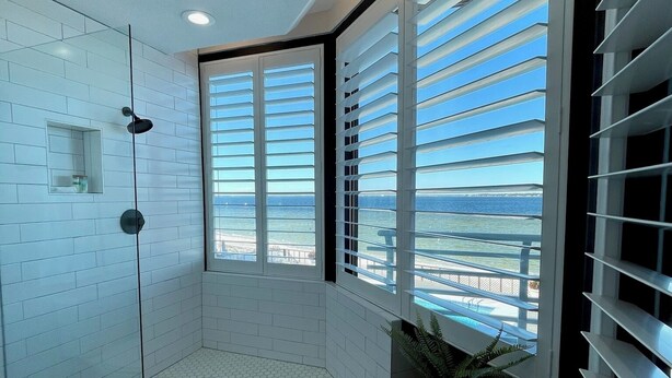 A modern bathroom with a glass-enclosed shower, white tiled walls, and a view of the ocean through large windows with blinds.