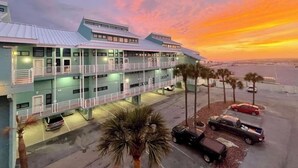 A multi-story hotel with balconies, parking, and a view of the ocean at sunset.