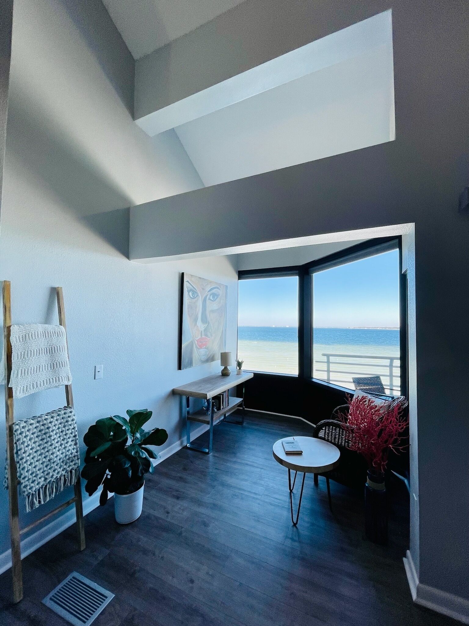 A modern room with a large window overlooking a beach, a wooden floor, a small round table, and a painting on the wall.