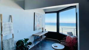 A modern room with a large window overlooking a beach, a wooden floor, a small round table, and a painting on the wall.