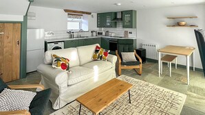 Un salon moderne avec un canapé, une table basse et des fauteuils. On aperçoit, au fond, une cuisine avec des meubles verts et une fenêtre.
