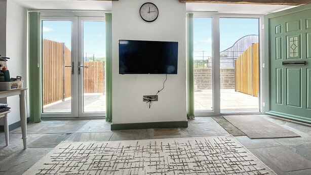 Une pièce dotée d’un téléviseur à écran plat fixé au mur, d’une horloge et d’une porte donnant sur un espace extérieur délimité par une clôture en bois.