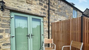 Un bâtiment en pierre avec une porte verte, une clôture en bois et deux chaises blanches accompagnées de petites tables en bois.
