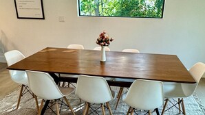 Ein Konferenzraum mit einem großen Tisch, weißen Stühlen, einer Blumenvase, einem gerahmten Bild und einem Fenster mit Blick auf Bäume.