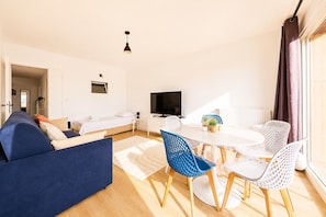A modern living room with a sofa, a dining table with chairs, a TV, and a window with curtains.