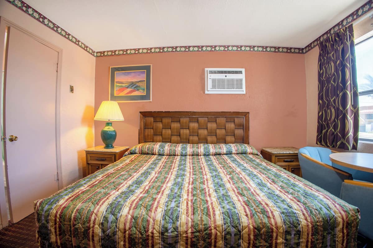 A hotel room with a large bed, a bedside table with a lamp, a small table with a chair, and a window with curtains.
