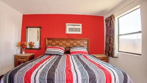 A hotel room with a red wall, a wooden headboard, a large bed with striped bedding, a nightstand with a vase of flowers, and a window with curtains. - Sands Motel By OYO El Paso Near Airport (El Paso)
