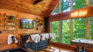 Ein gemütliches Zimmer mit Holzwänden, einem Billardtisch, einer Bar mit Hockern und einem großen Fenster mit Blick auf den Wald.