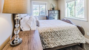A bedroom with a bed, a wooden nightstand with a lamp, a dresser, and a window with a view of trees. - 30% OFF PROMO! East Ogden Dream, Newly Built, BBQ, Patio, Views, Dedicated Spa! (Ogden)