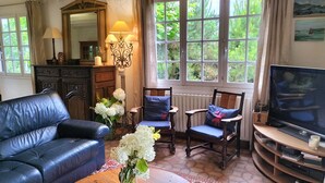 Un salon avec un canapé en cuir noir, deux fauteuils en bois avec des coussins, une table sur laquelle est posé un vase de fleurs, un téléviseur et une grande fenêtre donnant sur un paysage verdoyant.