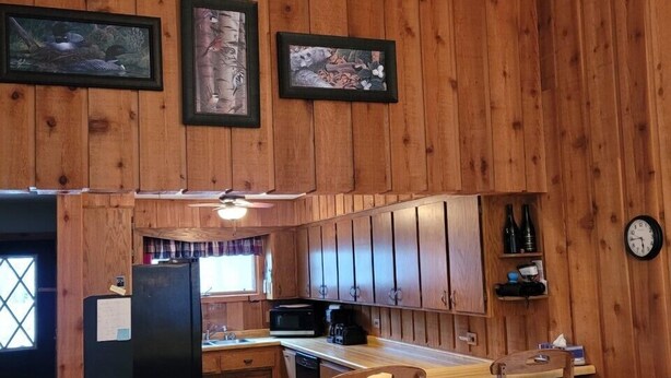 A wooden cabin interior with a kitchen area featuring a bar with stools, a ceiling fan, and framed artwork on the walls. - Cavalier #2 (Cable)