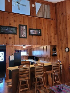 Het interieur van een houten chalet met een keukenhoek, inclusief een bar met krukken, een plafondventilator en lijstwerken aan de wanden.