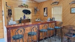 A wooden-paneled room with a bar area featuring four bar stools and a wooden table. - The Lodge (Cable)