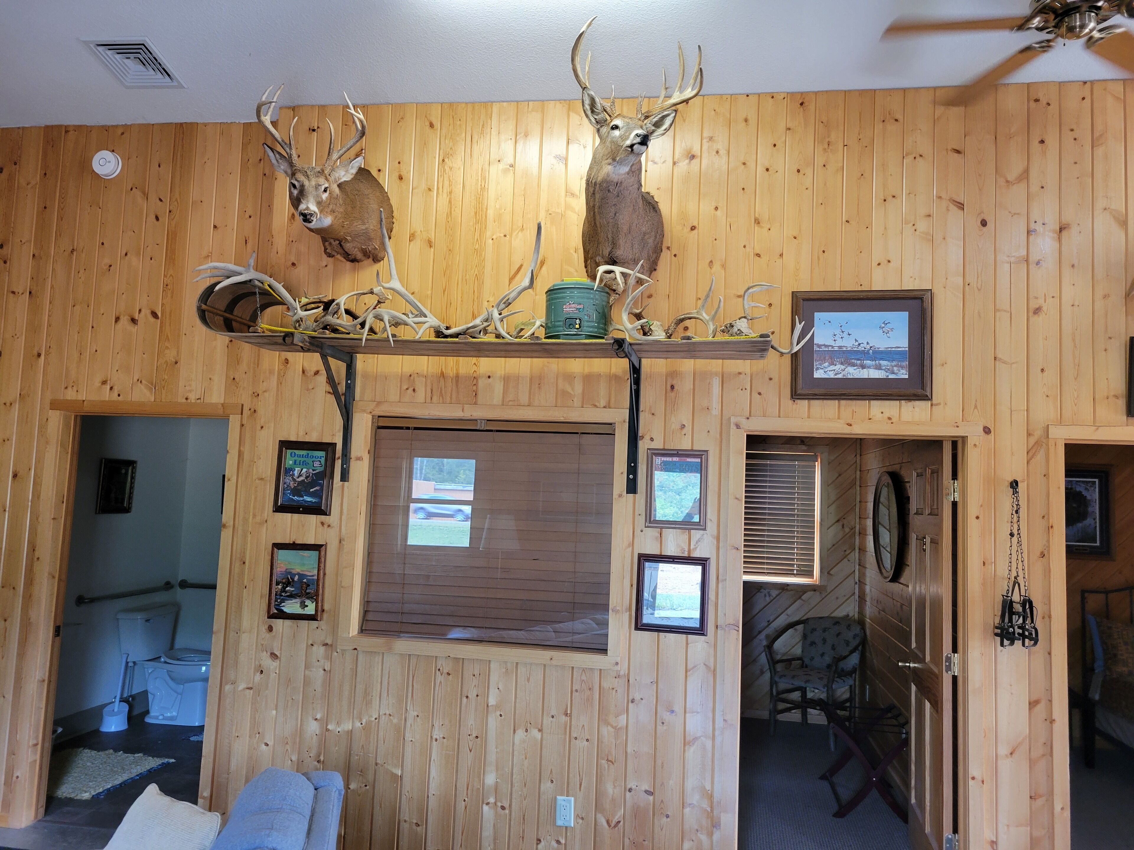A wooden interior with a mounted deer head, framed pictures, and a window with blinds.