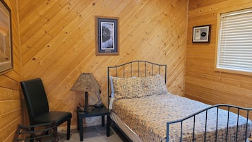 Habitación con paneles de madera, una cama, una silla, una mesita de noche y una ventana con persianas.