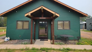 A green cabin with a 'Cabin For Rent' sign, a front door, and a small patio with a table and chairs. - The Lodge (Cable)