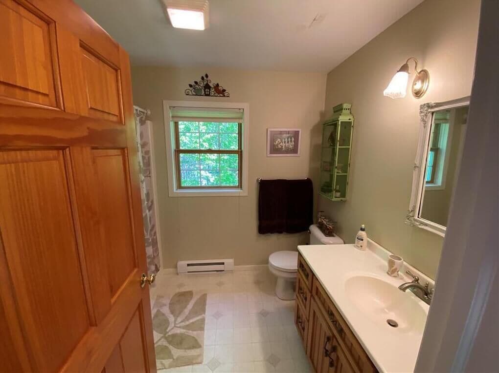 Un baño con puerta de madera, ventana, inodoro, lavamanos y espejo.