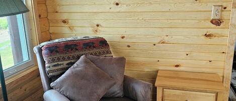 A cozy room with a wooden log wall, a brown armchair with a patterned throw, a small wooden nightstand, and a lamp.