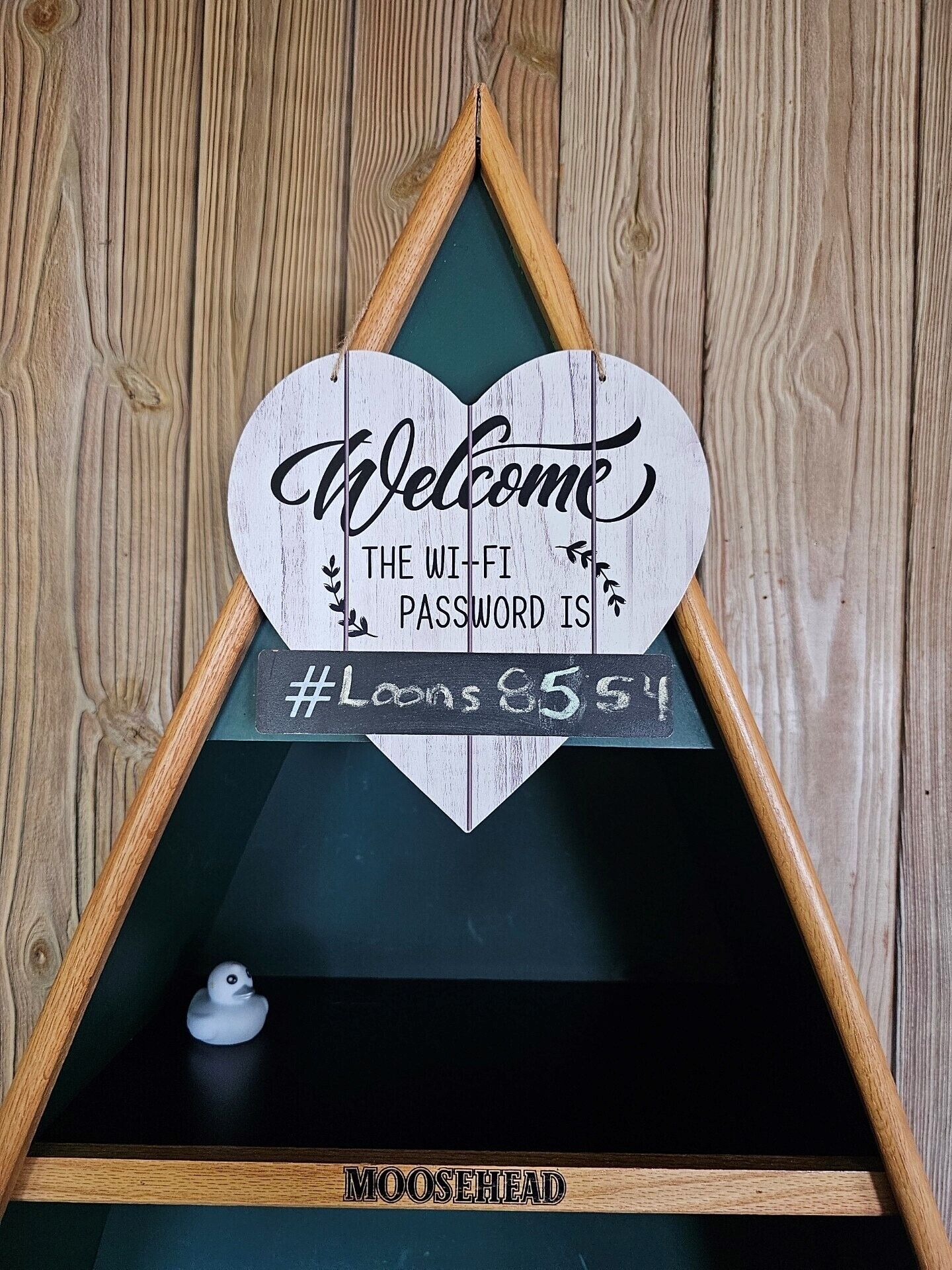 Una estantería de madera triangular con un letrero con forma de corazón que dice “Bienvenidos, la contraseña del Wi-Fi es #Loons 8551” y un patito de goma.