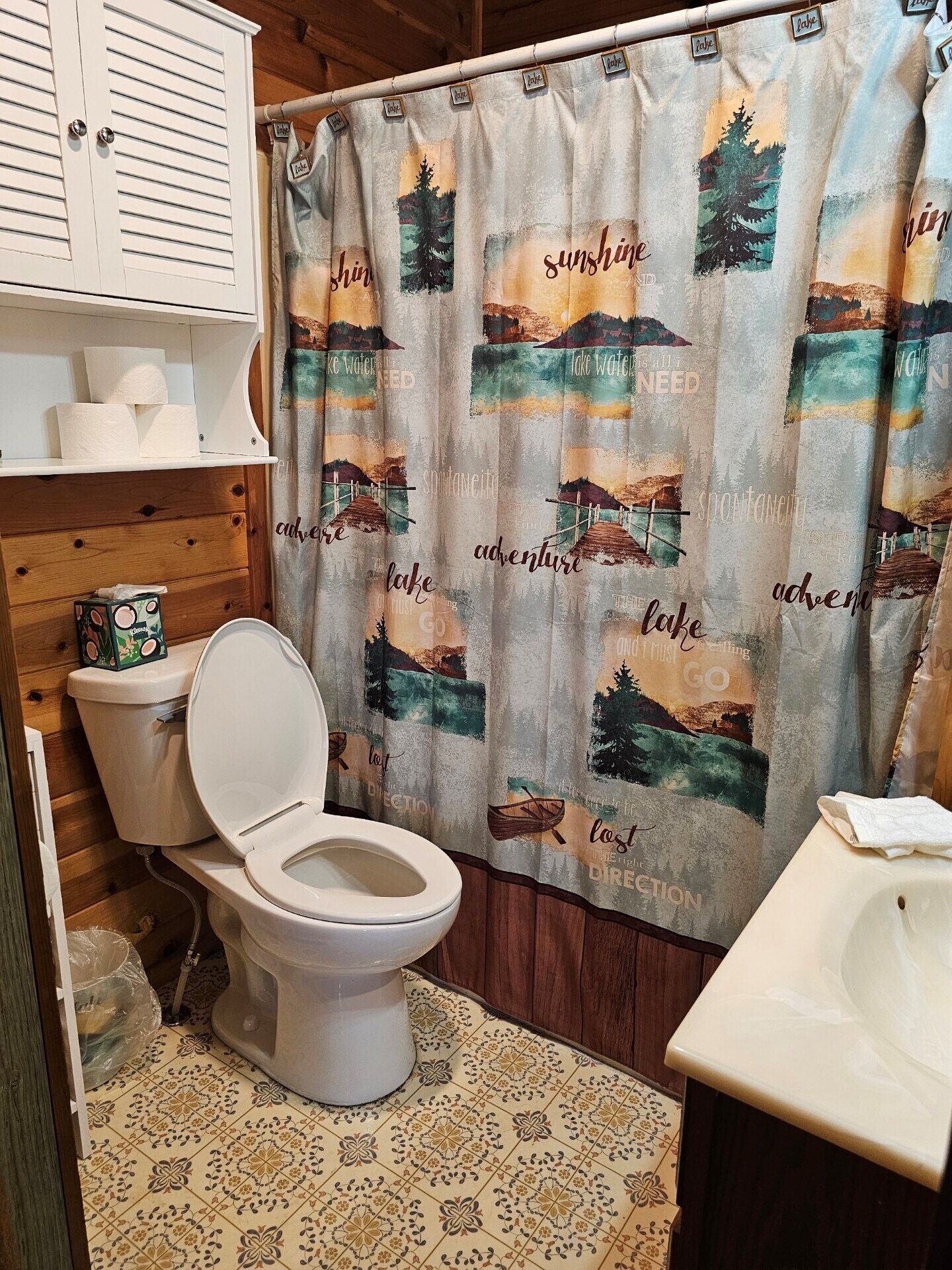 Ein Badezimmer mit Toilette, einer Duschtasse und einem Waschbecken. Der Duschvorhang zeigt ein Naturmotiv, der Boden ist gemustert.