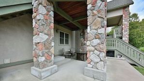 A covered outdoor patio area with stone pillars, a wooden ceiling, and a staircase leading to a second floor. - Unit 109 2 Bdrm/ 2 BA (Birchwood)