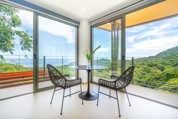 Un balcón con una mesa redonda y dos sillas, con vista a un paisaje verde y exuberante y el mar.