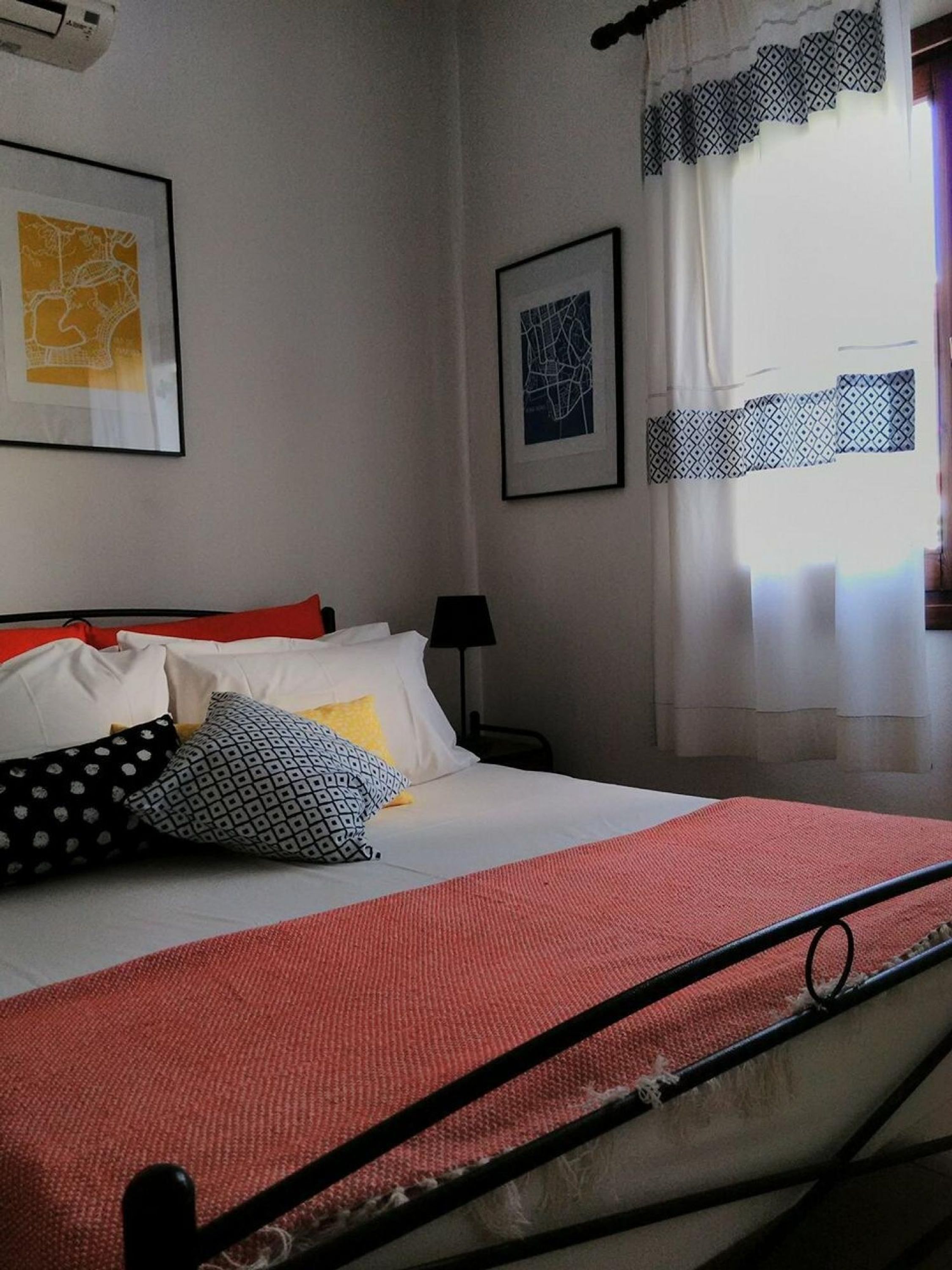 A bed with a red blanket, white pillows, and patterned pillows. There are two framed artworks on the wall, a map and a geometric design. A lamp is on the nightstand beside the bed.