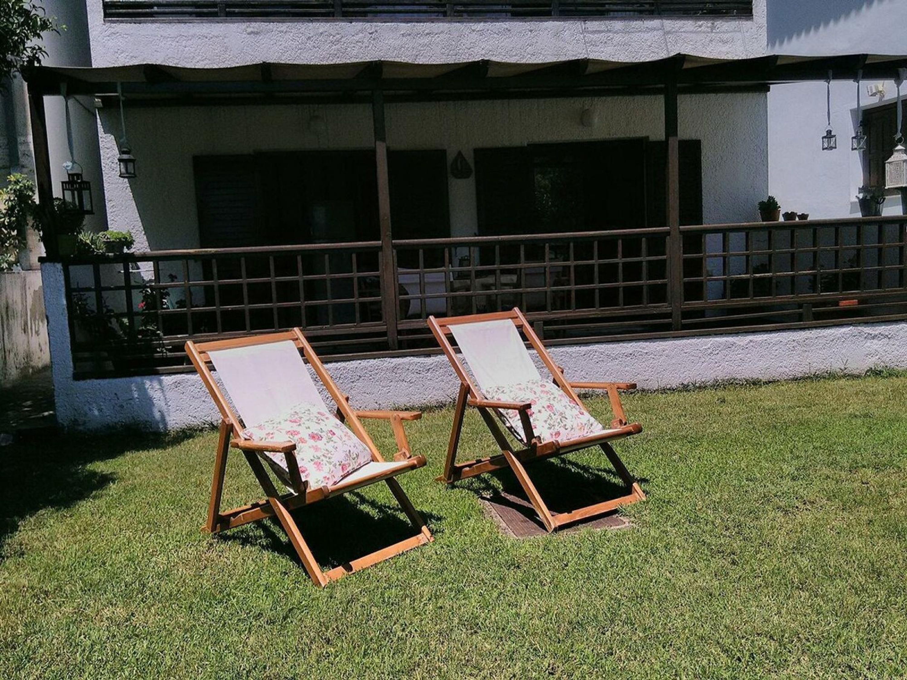 Dos sillones de madera con cojines estampados sobre un césped frente a un edificio de fachada blanca y barandilla marrón oscuro.