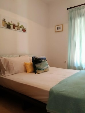 A bedroom with a bed, pillows, a shelf with potted plants, a framed picture, and a window with curtains.