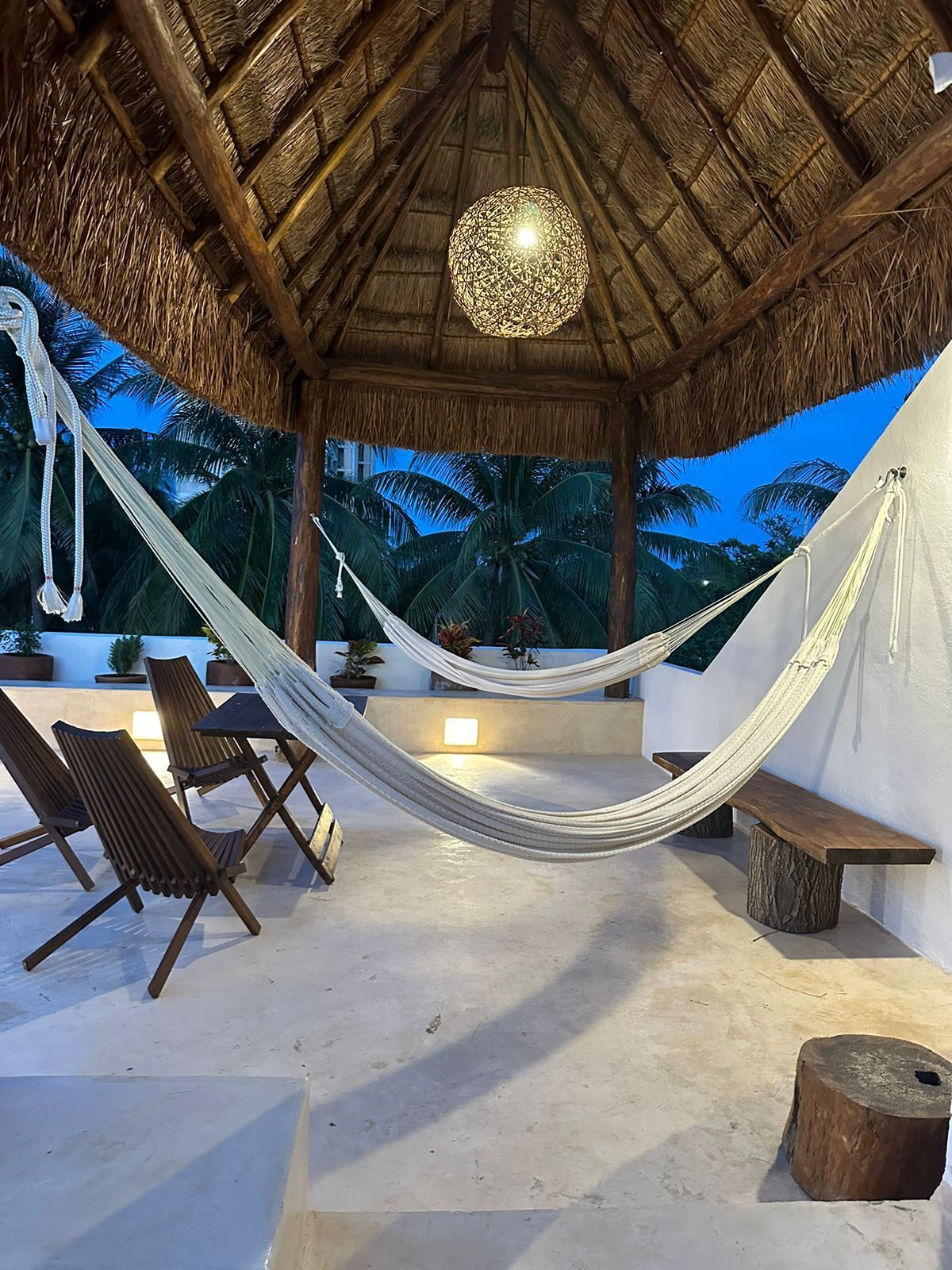 A hammock under a thatched roof, wooden chairs, and a bench in a tropical setting.